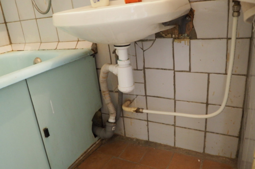 Состояние старого водопровода из разного вида труб и старая, неаккуратная плитка на полу и стенах в ванной под умывальником
